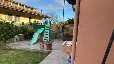 A small backyard features a green and red slide, potted plants, tiled patio, grassy area, some trees, a wooden fence, and a yellow house in the background.