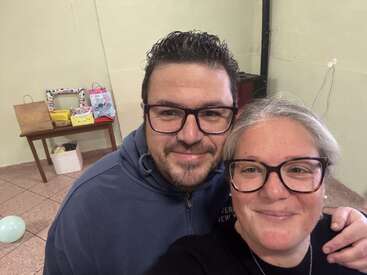 A smiling man and woman wearing glasses pose for a selfie indoors. Behind them, there's a table with gift bags and presents, and a balloon on the floor.