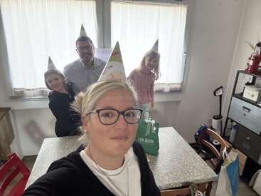 Una familia de cuatro miembros, todos con gorros de fiesta, sonríe junta en una acogedora cocina. Sobre la mesa hay bolsas de regalo; están celebrando un cumpleaños o una ocasión especial.