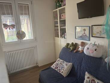 Une chambre d'enfant chaleureuse, avec un canapé bleu, un coussin décoratif, des peluches et une poupée. Une étagère, des dessins et des jeux agrémentent cet espace lumineux et baigné de soleil.