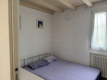 A small, minimalist bedroom with a simple metal bed frame, light purple bedding, wooden ceiling beams, a curtained window, and a single framed artwork on the wall.
