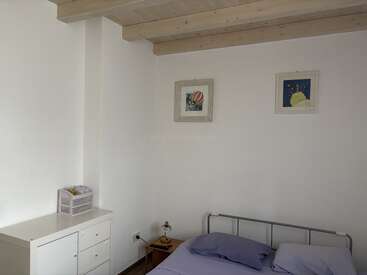 This image shows a minimalist bedroom with white walls, a metal bed frame, pastel bedding, two framed artworks, a small dresser, and a wooden ceiling with exposed beams.