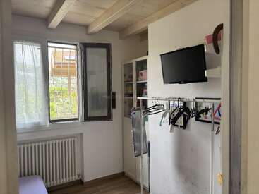 This small room has a window, radiator, a wooden ceiling, clothes hangers, a television mounted on the wall, shelves, and a cabinet with various items.