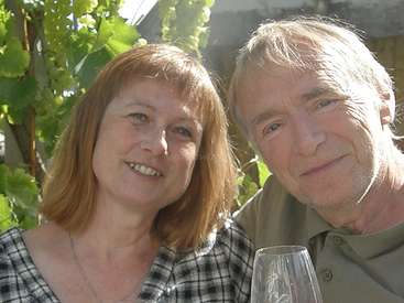Das Bild zeigt ein Paar mittleren Alters, das für ein Foto posiert. Die Frau auf der linken und der Mann auf der rechten Seite lächeln und schauen in die Kamera.