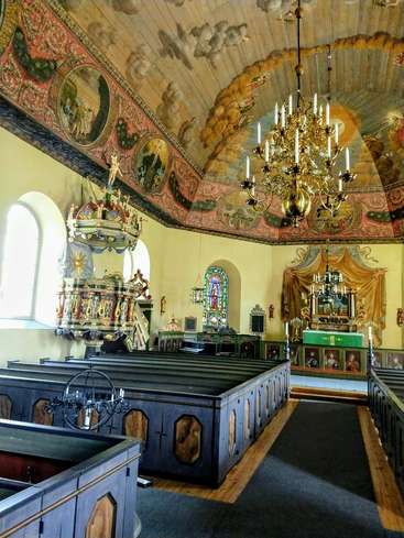 Das Bild zeigt das Innere einer Kirche mit kunstvollen Verzierungen, einem großen Kronleuchter und Kirchenbänken, während im Hintergrund ein Glasfenster und ein Altar zu sehen sind.