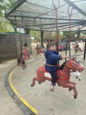 Un jeune garçon monte sur un cheval de manège rouge sous une verrière. D'autres enfants s'amusent eux aussi sur ce manège d'époque, entouré d'équipements de jeux en plein air et d'arbres.