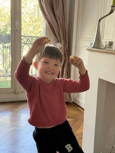 Un jeune enfant, le sourire aux lèvres, se tient debout à l'intérieur, tenant dans ses deux mains de petits objets ronds. La lumière du soleil pénètre à flots par les grandes fenêtres, créant une atmosphère chaleureuse et joyeuse.