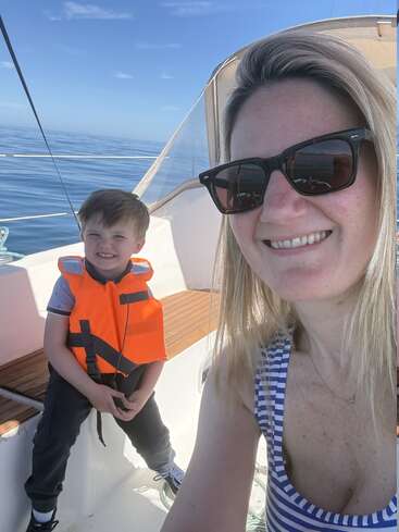 Une femme et un jeune garçon sourient sur un bateau. Le garçon porte un gilet de sauvetage orange. Il fait beau, et la mer est calme et bleue.