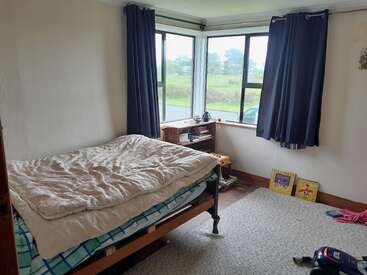 This cozy bedroom features an unmade bed, bay windows with dark curtains, a small wooden desk, light walls, books, and a rug on the floor.