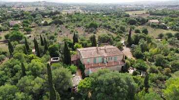 Esta imagen muestra una gran casa de campo rodeada de vegetación densa y frondosa y árboles altos, situada en un vasto paisaje rural con colinas ondulantes más allá.