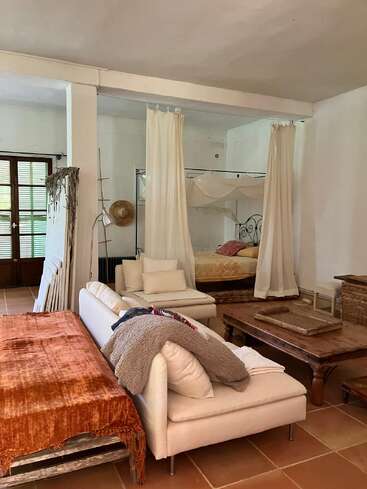 Este acogedor salón cuenta con un sofá blanco, una mesa de centro rústica, un sofá-cama con una manta naranja, una cama con dosel y cortinas, y luz natural procedente de las puertas francesas.