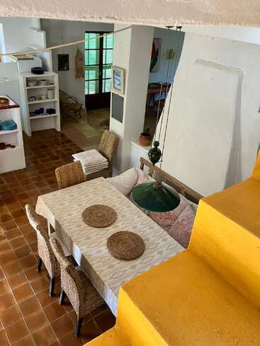 Una acogedora zona de comedor con sillas de mimbre, un banco acolchado, escaleras amarillas y un mantel estampado. Las estanterías abiertas, la lámpara verde y la luz natural aumentan la calidez.