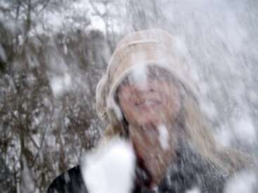 Uma pessoa usando um chapéu de inverno bege está do lado de fora, parcialmente escondida por uma rajada de neve que cai, com um fundo invernal desfocado, com árvores sem folhas.