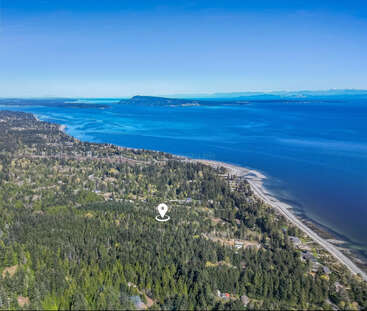 Vista aérea de una exuberante zona costera boscosa con un litoral sinuoso, casas residenciales, un océano azul extenso, islas lejanas y un cielo azul despejado. Se muestra el marcador de ubicación.