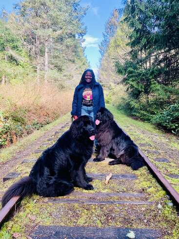 Una mujer sonríe de pie sobre las viejas vías del tren en un bosque, acompañada de dos grandes perros negros sentados alegremente, rodeados de frondosos árboles verdes.