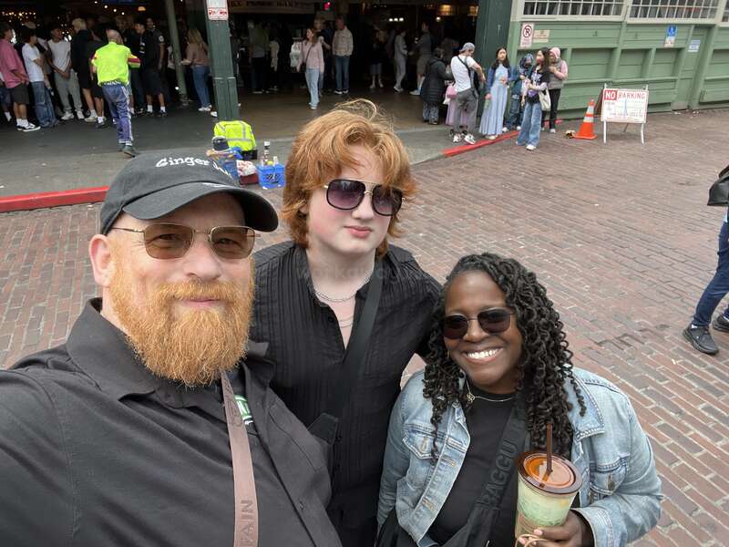 Tres amigos posan para un alegre selfie en una calle de ladrillo, con gafas de sol y ropa informal. Una multitud y la actividad del mercado suceden en el fondo.
