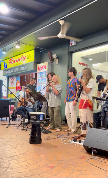 Un groupe de musiciens se produit en direct devant le Ramly Café le soir, chantant et jouant de leurs instruments. Un pot à pourboires est placé devant le groupe.
