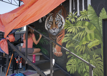 Une femme est assise sur des marches, à l'abri d'une bâche orange, et peint sur un mur sombre une grande fresque réaliste représentant un tigre entouré de feuilles de jungle d'un vert luxuriant.