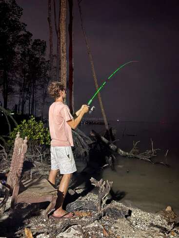 Un homme en tenue décontractée se tient près d'une rive, la nuit, tenant une canne à pêche lumineuse, éclairée par une source de lumière, avec des arbres et une vieille chaise à ses côtés.