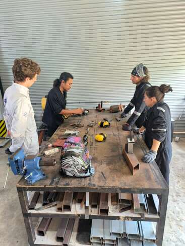 Quatre personnes sont debout autour d'une table d'atelier recouverte d'outils et de casques de soudure. Elles semblent concentrées, peut-être en train de recevoir des instructions ou de discuter ensemble d'un projet de métallurgie à l'intérieur d'un atelier.