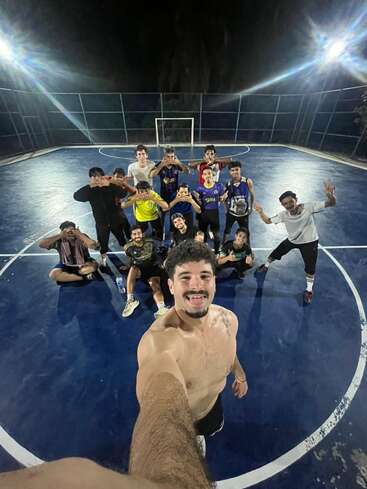 Un groupe d'amis pose pour un selfie sur un terrain de futsal bleu en plein air, de nuit, souriants et enjoués après un match endiablé sous les projecteurs.