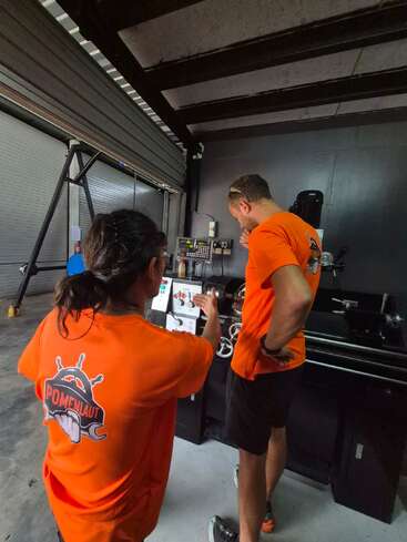 Deux personnes vêtues de chemises orange travaillent dans un atelier. L'une d'elles consulte le mode d'emploi d'une machine tandis que l'autre l'observe. Sur leurs chemises figure un logo portant l'inscription « Pomenlaut ».
