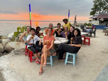 Un groupe d'amis prend un verre dans un café en bord de mer au coucher du soleil, assis autour d'une table et souriants, avec l'océan et un ciel coloré en arrière-plan.