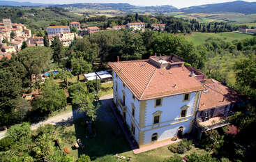 This image shows a large villa with a red-tiled roof surrounded by lush greenery, trees, and gardens, set in a scenic countryside village with rolling hills.
