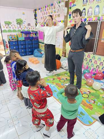 In a colorful classroom, two teachers lead a group of young children in an interactive activity, with everyone touching their heads and following instructions enthusiastically.