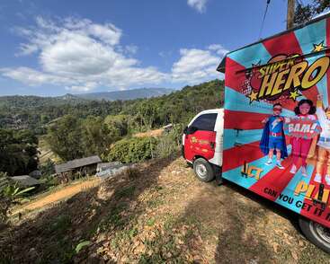 A colorful van with "Super Hero" graphics is parked on a hill overlooking a scenic landscape of mountains, trees, village houses, and a bright blue sky.