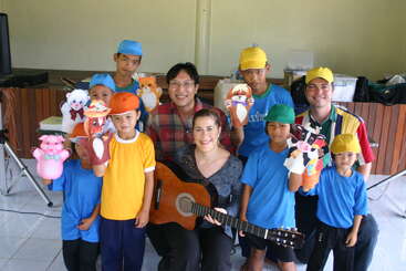 The image shows a group of children and adults posing together, with the children holding paper puppets and one woman holding a guitar.