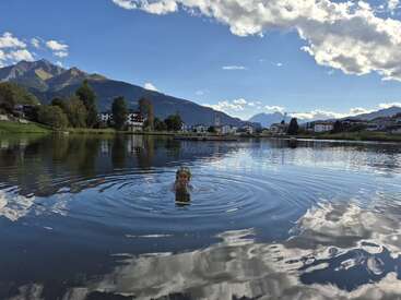A person swims alone in a tranquil lake surrounded by mountains, trees, and a small town under a blue sky with scattered clouds, reflecting peaceful serenity.