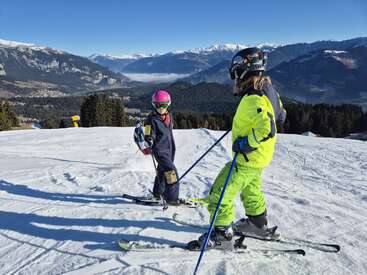 Two skiers, dressed in colorful winter gear, stand on a snowy mountain slope. Majestic mountains, forests, and a clear blue sky create a breathtaking backdrop.