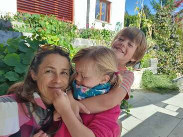 Das Bild zeigt eine Frau und zwei Kinder, die an einem sonnigen Tag vor einem Haus mit Garten für ein Foto posieren.