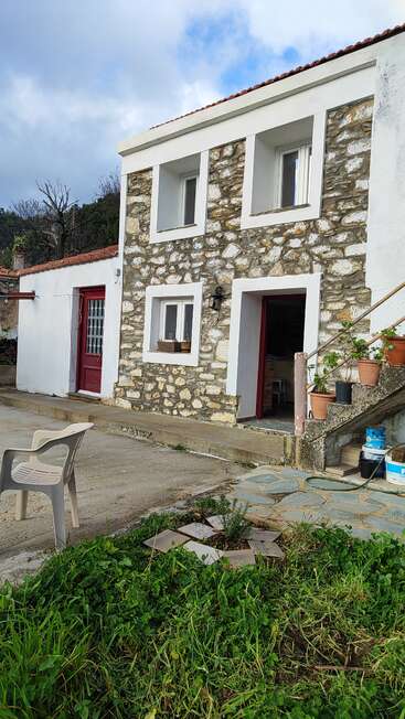 Uma casa rústica de pedra com detalhes em branco, portas vermelhas e vasos de plantas nas escadas. Tem uma cadeira de plástico, vegetação e um céu azul nublado lá em cima.
