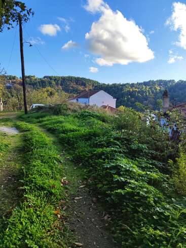 Uma trilha estreita de terra, ladeada por uma grama verdejante, leva a casas brancas, aninhadas entre colinas e árvores, sob um céu azul brilhante com nuvens.
