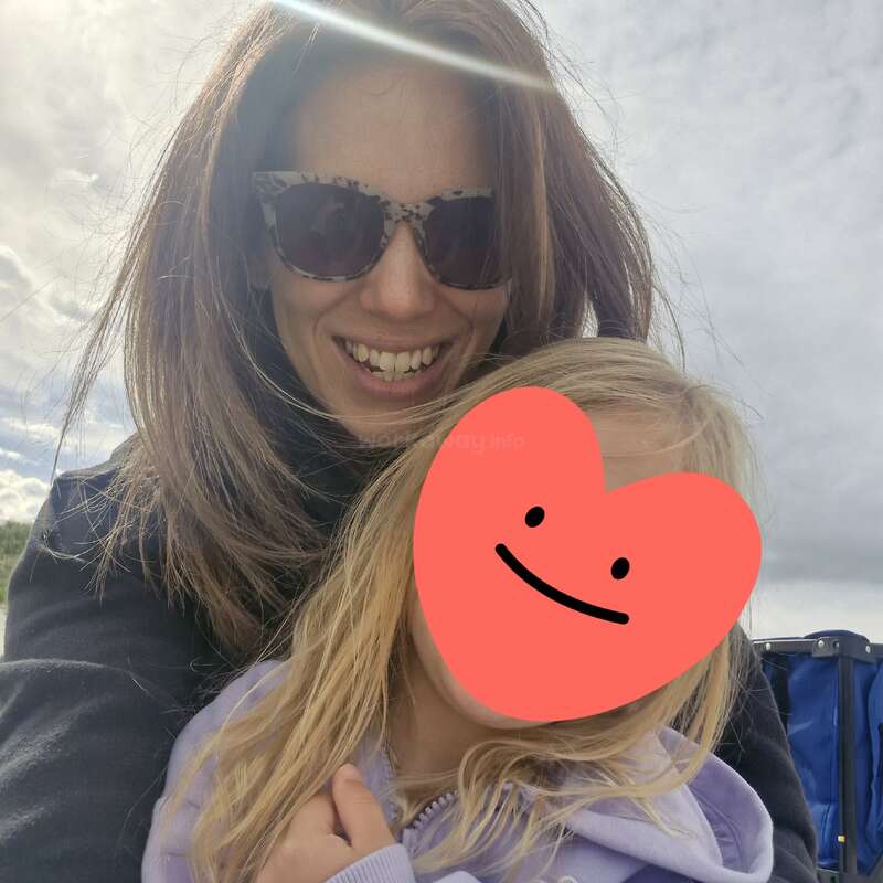 Una mujer sonriente que lleva gafas de sol abraza a un niño pequeño de pelo rubio. La cara del niño está tapada por una pegatina roja en forma de corazón con una carita sonriente.