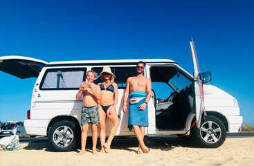 Three people in swimwear stand smiling beside a white van at the beach, under a clear blue sky, enjoying a sunny, relaxed vacation moment together.