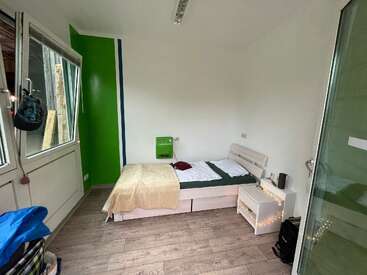 A imagem mostra um pequeno quarto com uma cama, uma mesa de cabeceira e um armário, com paredes brancas, piso de madeira e uma janela, com uma parede verde de destaque no lado esquerdo.