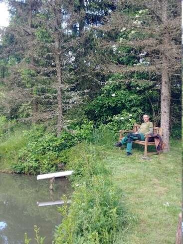 Un homme est assis sur un banc en bois dans un cadre paisible et verdoyant, au bord d'un étang calme, entouré d'herbe, de buissons et de grands arbres à feuilles persistantes.