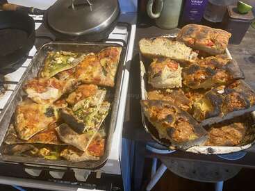 Duas bandejas sobre o fogão da cozinha exibem fatias de pizza caseira de vários sabores e pão focaccia rústico, algumas com queijo e vegetais à vista e crostas douradas e levemente tostadas.
