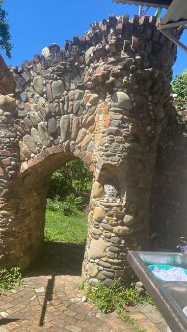 Esta imagem mostra um arco rústico de pedra com tijolos, um nicho com uma escultura decorativa de um leão, vegetação, um céu azul claro e uma pia de metal ao ar livre.