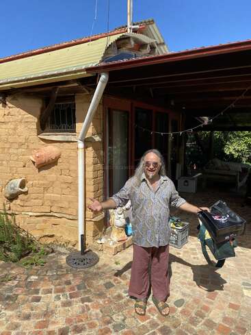 Uma pessoa de cabelo comprido e óculos escuros está em um caminho de tijolos do lado de fora de uma casa rústica, sorrindo, segurando sacolas, cercada por um jardim e objetos decorativos sob a luz do sol.