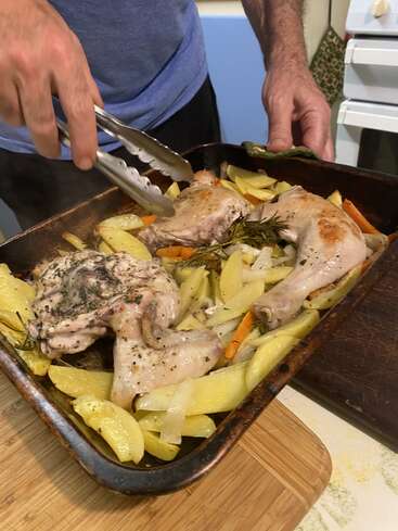 Uma pessoa está preparando um prato assado com coxinhas de frango, batatas, cenouras e ervas em uma assadeira, usando uma pinça para servir a refeição.