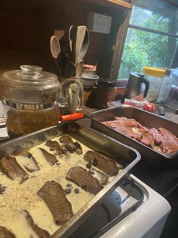 Esta cena aconchegante na cozinha mostra um pudim de pão embebido em creme e uma bandeja com carne temperada, com utensílios, um bule de chá e vegetação visíveis do lado de fora da janela.