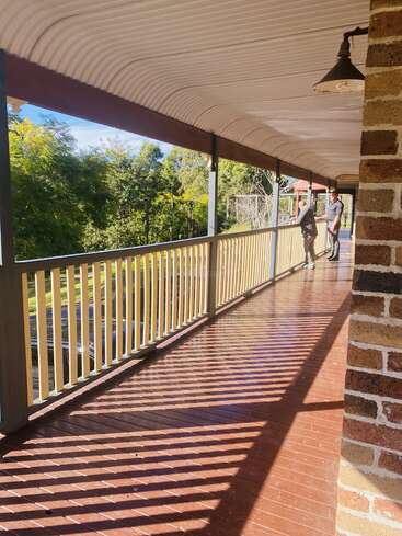 L'image représente une allée couverte avec une balustrade et un pilier en briques, avec un homme debout sur le côté droit, sur fond d'arbres et de ciel bleu.