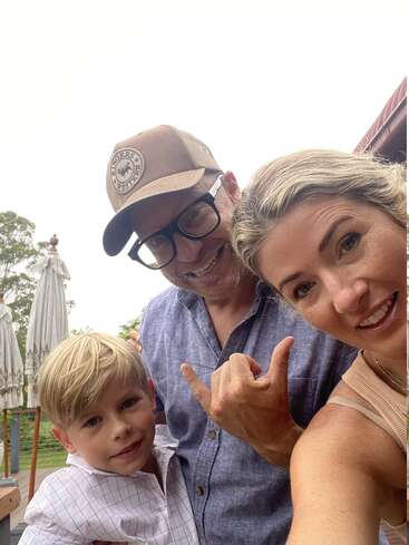 L'image montre une famille de trois personnes posant pour un selfie, le père portant une casquette de baseball et la mère et l'enfant faisant un geste de la main.