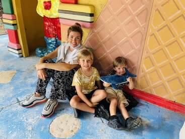 L'image représente une femme et deux jeunes garçons assis par terre devant un mur coloré, la femme portant une chemise blanche et un pantalon noir.