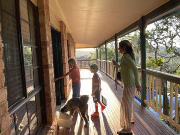 L'image représente une femme et deux enfants debout sous un porche, avec un chien, regardant à travers une porte moustiquaire une maison située de l'autre côté.