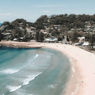 A imagem retrata uma cena de praia serena com uma pequena cidade aninhada nas colinas, com uma costa arenosa, ondas suaves e um cenário pitoresco de árvores e edifícios.